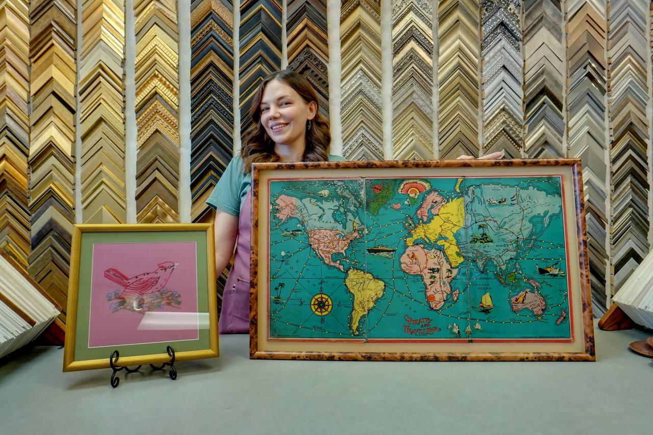 Jessica Edghill, third-generation owner of The Frame Up, smiling in front of the moulding corner-sample wall while holding a framed vintage world map, with a framed bird artwork on a display easel beside her